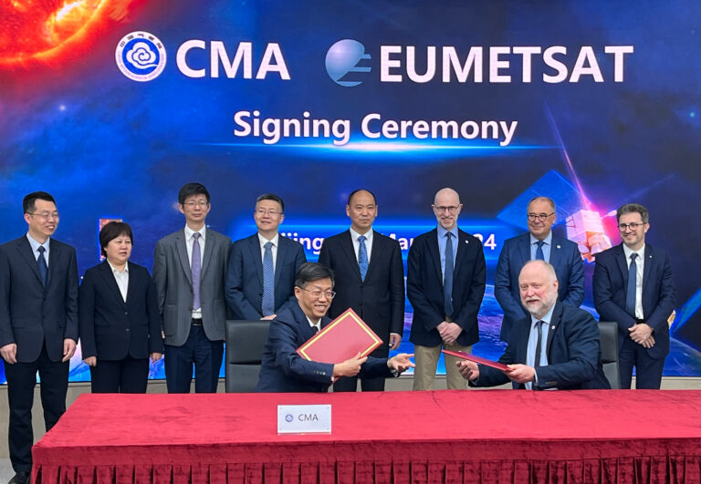 Zhenlin Chen (left), administrator of CMA, and Phil Evans (right), director-general of EUMETSAT, grin and hand each other signed partnership extension documents in front of a row of standing executives in suits and the word “Signing Ceremony”.