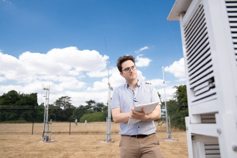 A researcher notes down readings from a weather station, in front of a blue sky.