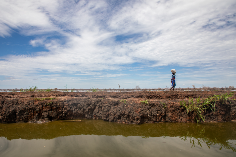 A woman walks carrying a bag on her head, on a mud path next to a body of water.
