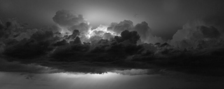 A black and white photo of gathered clouds with flashes of lightning.