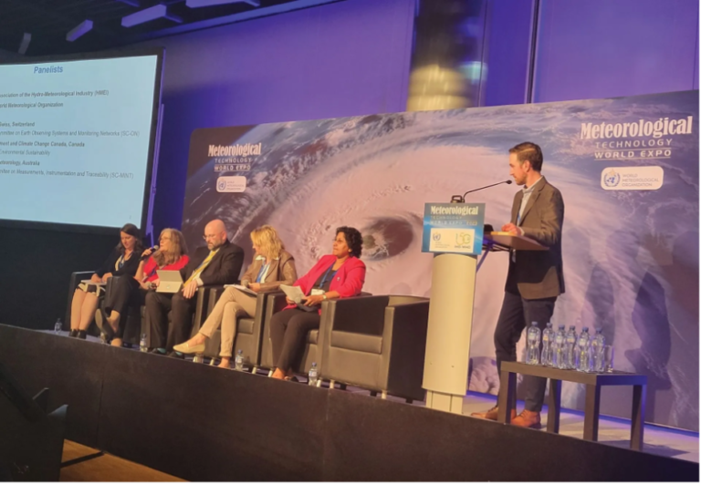 A panel of five experts sit in front of an aerial shot of a hurricane with the words "Meteorological Technology World Expo" on it.