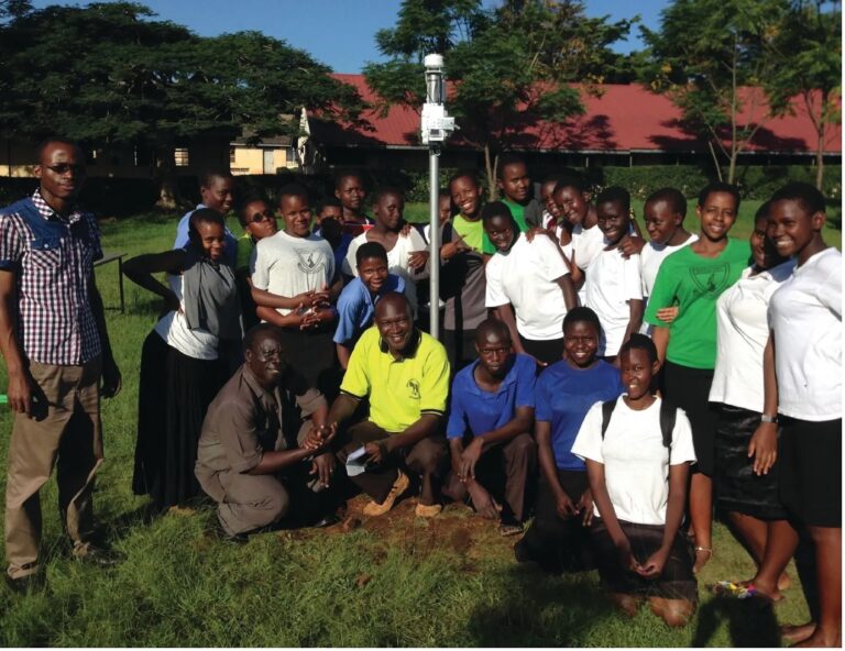 A group of adults and school children gather around a weather measurement station.
