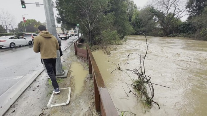 A man walks along a road, past a muddy overflowing river that has a tree poking out.