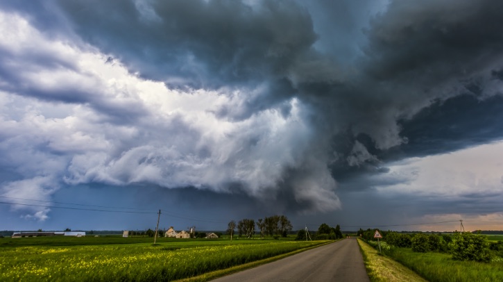 Dark storm clouds gather over a field, with a road running through the middle and green grass and crops on either side.
