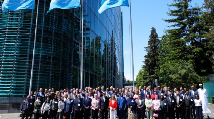 A large group of people pose for a photo outside a glass building with three United Nations flags, surrounded by trees under a clear sky.