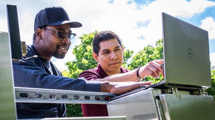 Two men work cheerfully at a laptop, with a blue sky and trees in the background.
