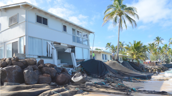 A white house is surrounded by large black rocks and debris washed up from the sea.