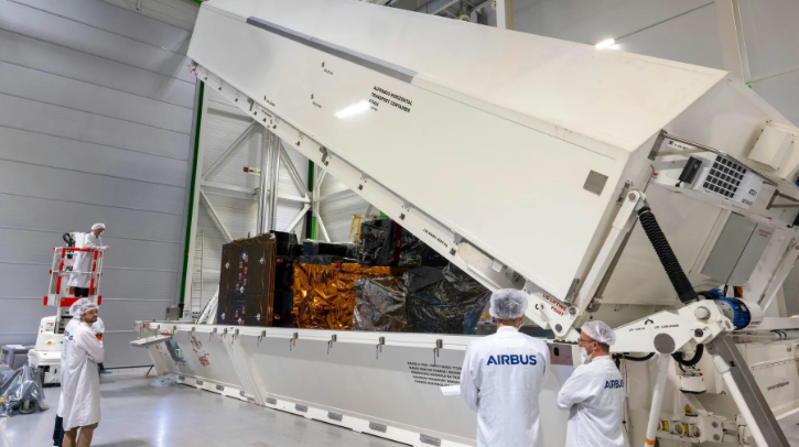 Technicians in labcoats with the word "Airbus" watch a satellite being packed.