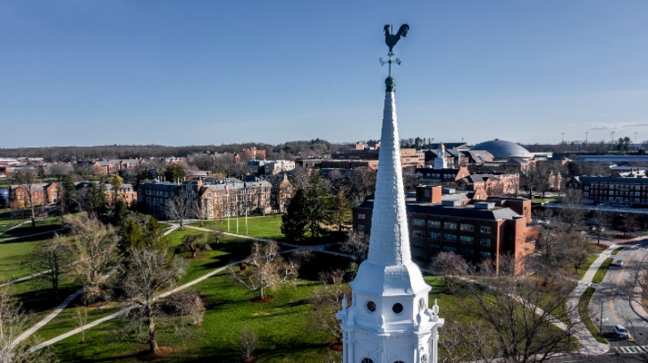 An aerial view of the University of Connecticut, with the spire in the foreground and green lawns behind among two and three-story brown-brick university buildings.