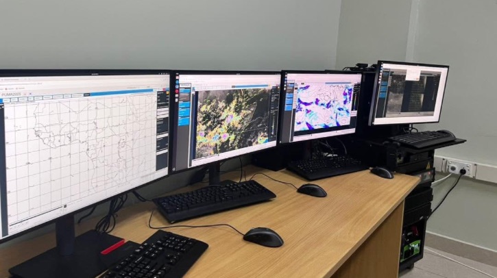 Four computer screens on a brown desk show maps and meteorological information.