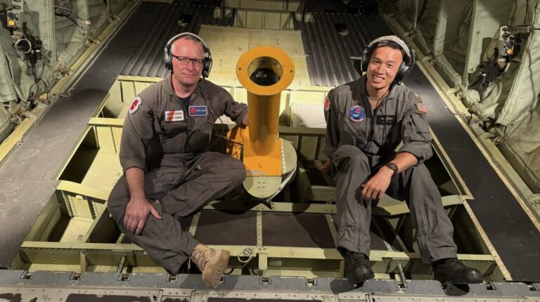Two scientists in work fatigues sit inside the cargo bay of a plane on a training mission.