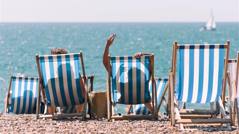 People sit in deck chairs on a pebble beach looking out to sea on a sunny day