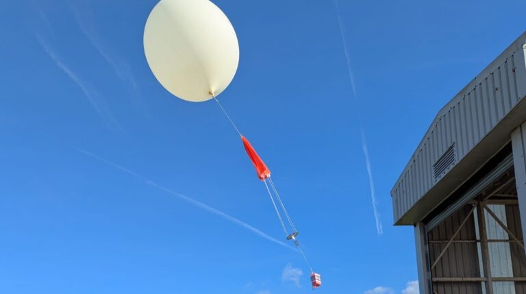 A large, white space weather balloon is launched into a clear blue sky to travel up through the atmosphere toward the stratosphere