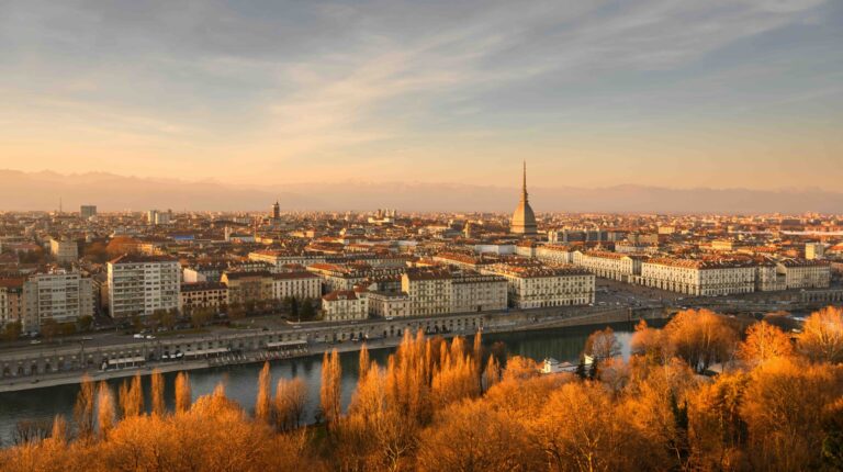 A skyline view of Turin at sunset in autumn, with a river in the foreground and a church spire in the mid-ground and a hazy blue-grey sky