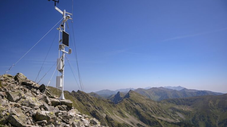 An automated, remote weather instrument on a rocky outcrop in a mountain range, with a clear blue sky above.