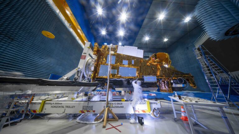 The MetOp-SG-B1 satellite – with white metal structure and bright gold pieces – undergoing tests in a large warehouse space, with a high ceiling a a scientists in the foreground in white overalls
